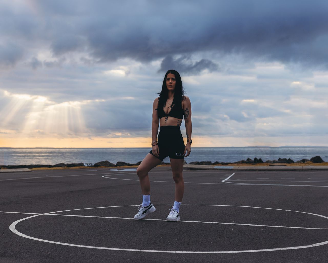 Athletic woman standing on a basketball court by the sea at sunset, representing strength and style.