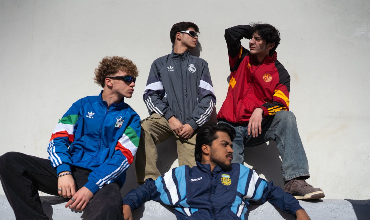 Four young men sitting against a wall, wearing sporty jackets and sunglasses.
