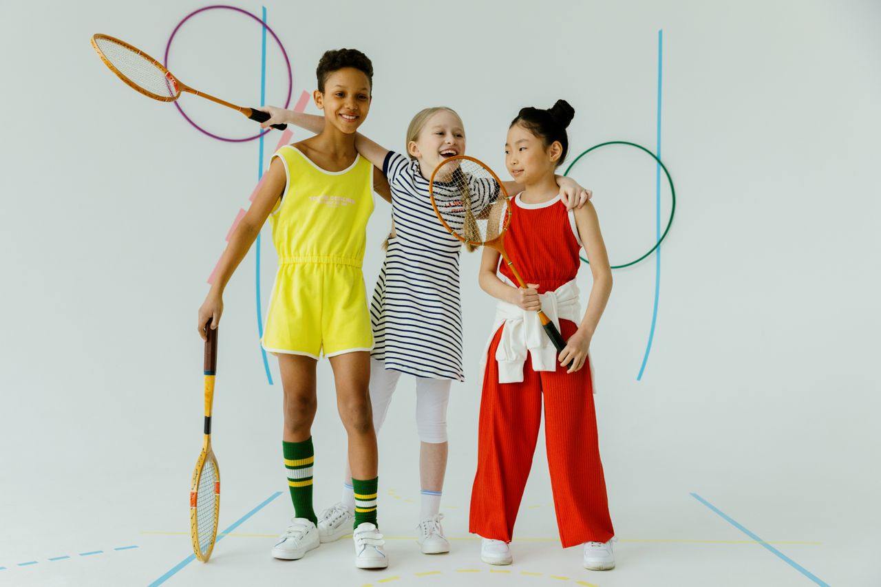 Three children in sportswear holding tennis rackets, smiling and embracing in a bright studio setting.
